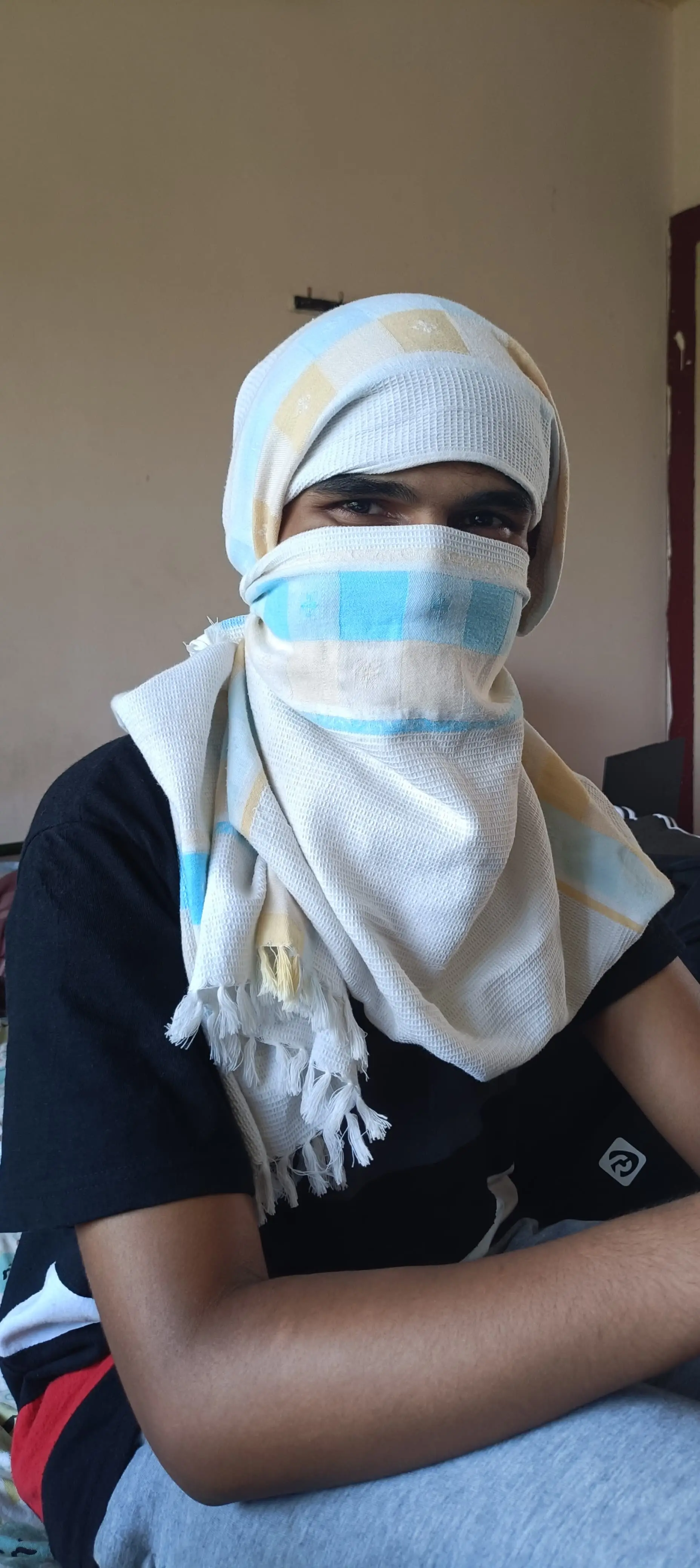 A close-up portrait of a person with their head and face wrapped in a checked cotton scarf, only the eyes visible.