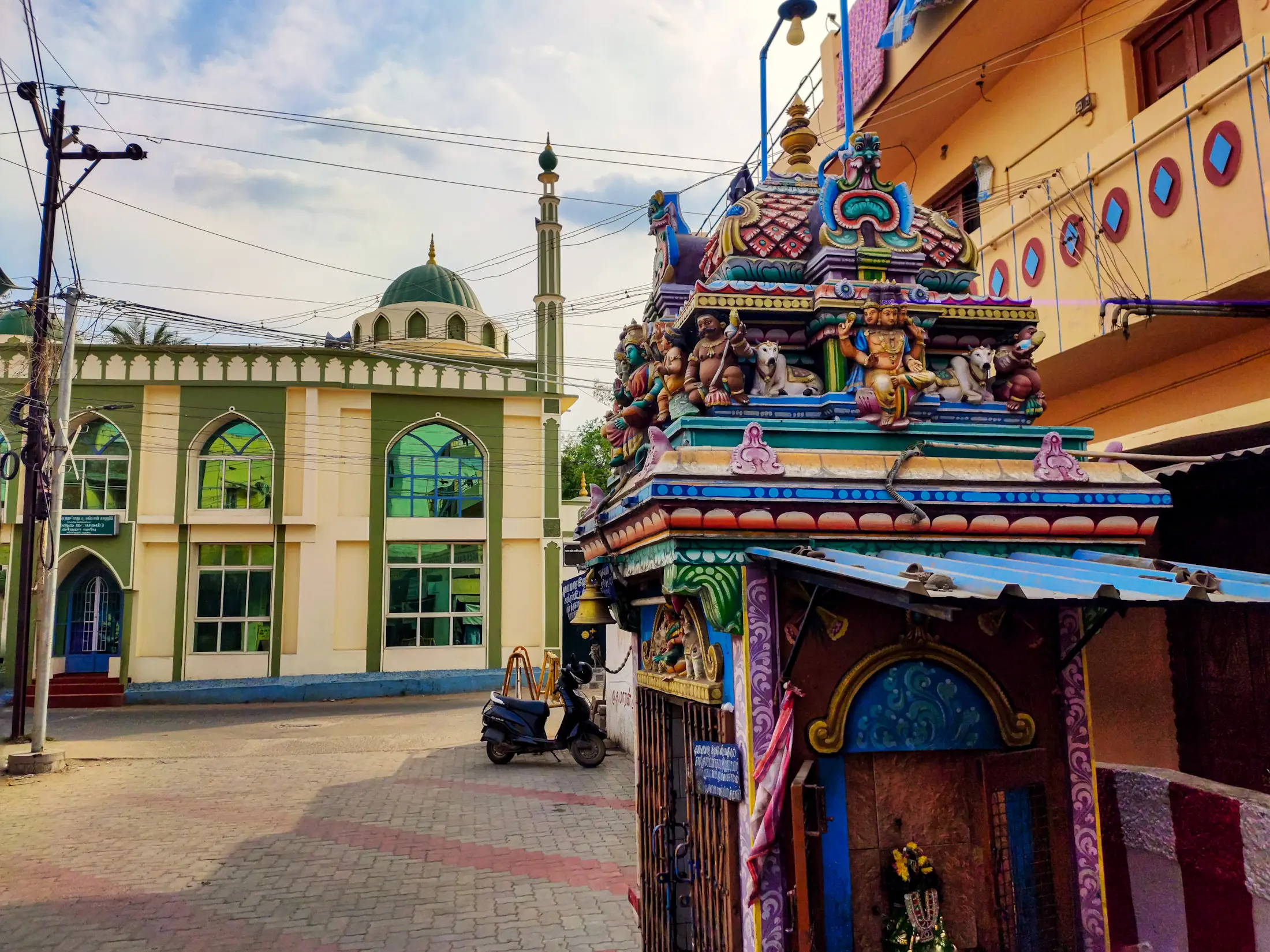 A small, brightly painted Hindu temple shrine roof in the foreground with a green-domed mosque and minaret visible down the street behind it.