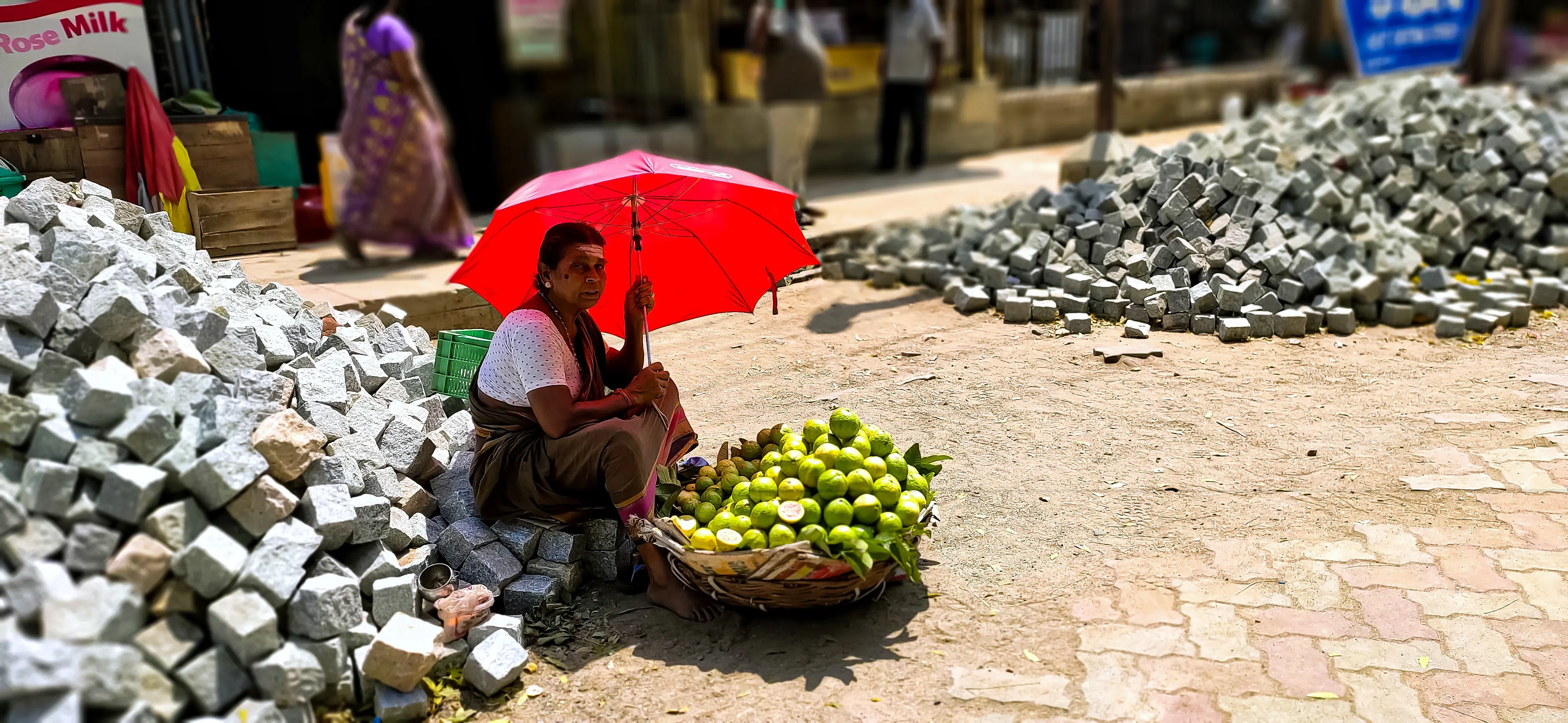 A street vendor sits beneath a bright red umbrella next to piles of granite cobblestones, a basket of limes in front of her.