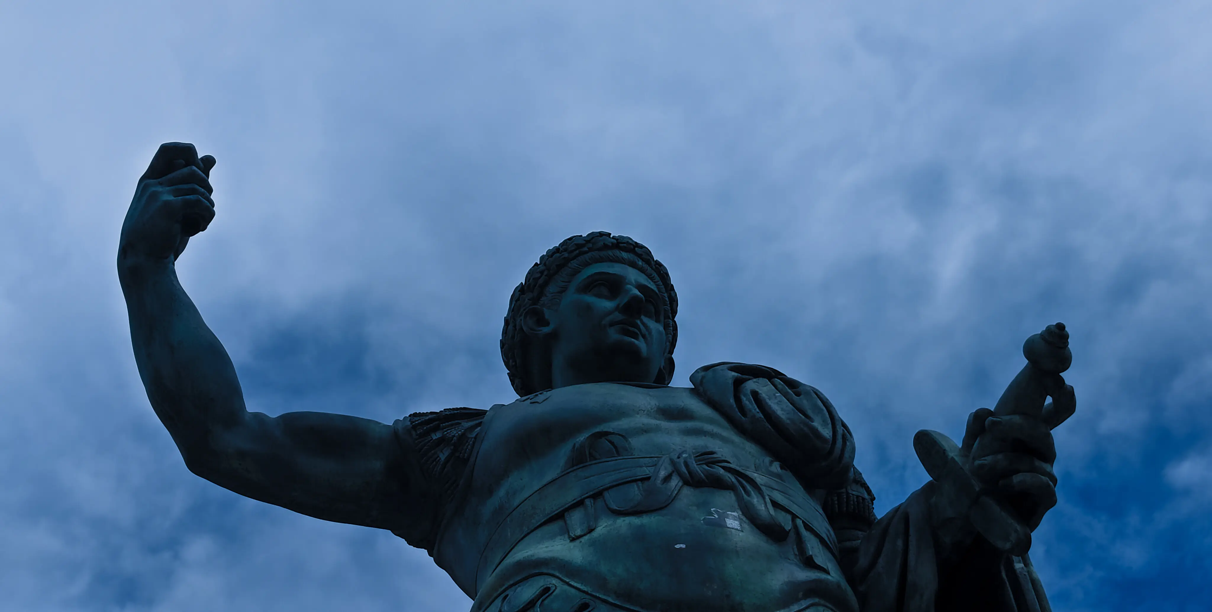 A weathered bronze statue of a robed, crowned figure raising one arm toward the sky, shot from below against a dusky, cloudy sky.