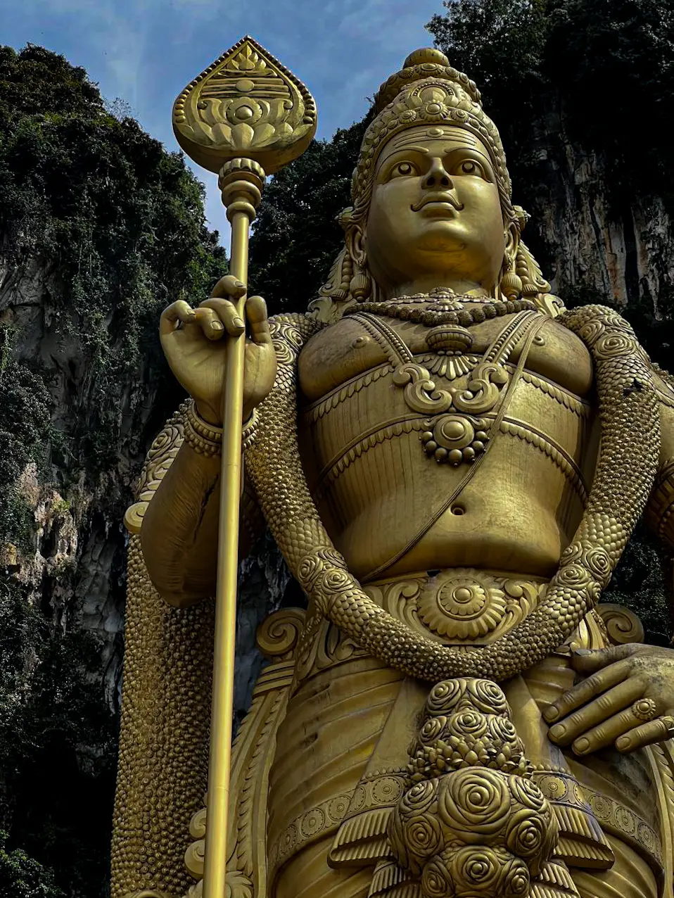 A towering golden statue of a deity holding a spear, set against a rock cliff face and greenery.