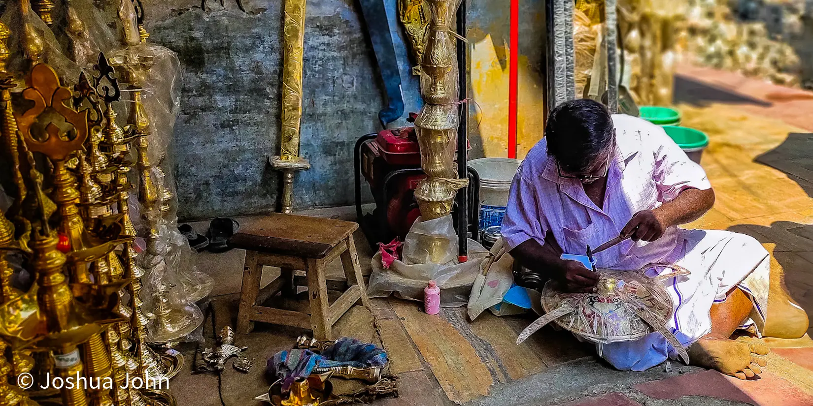 A craftsman sits on the ground polishing an ornate brass temple lamp, surrounded by rows of similar brassware.