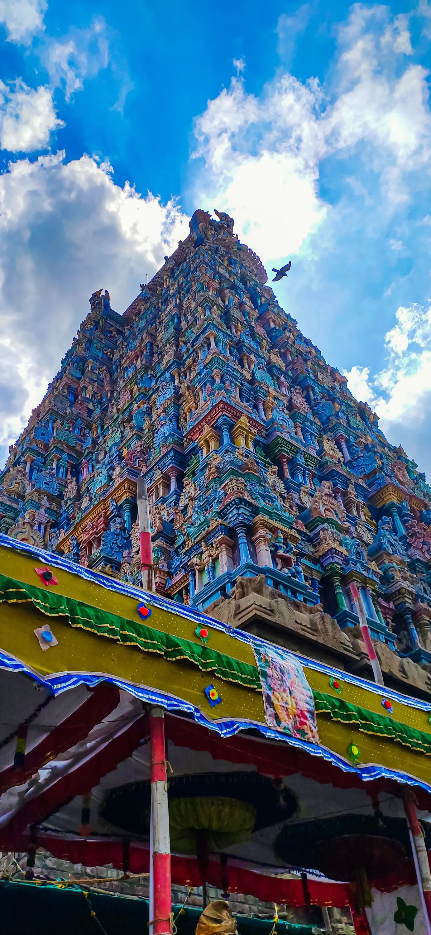 A tall, ornately carved South Indian temple gopuram tower rising against a bright blue sky with clouds, seen from below.