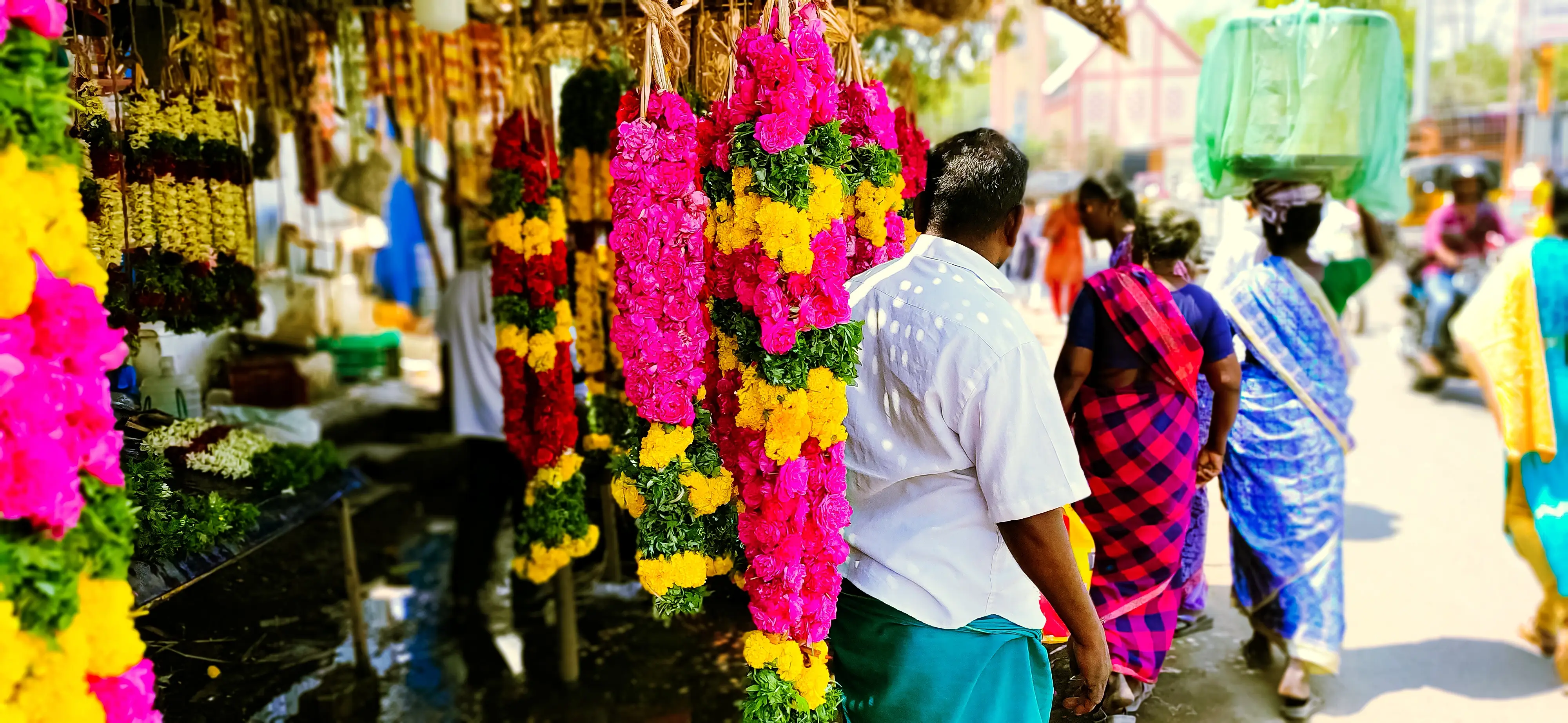 Strings of pink, yellow, and red flower garlands hang at a market stall as a man in a white shirt walks past.