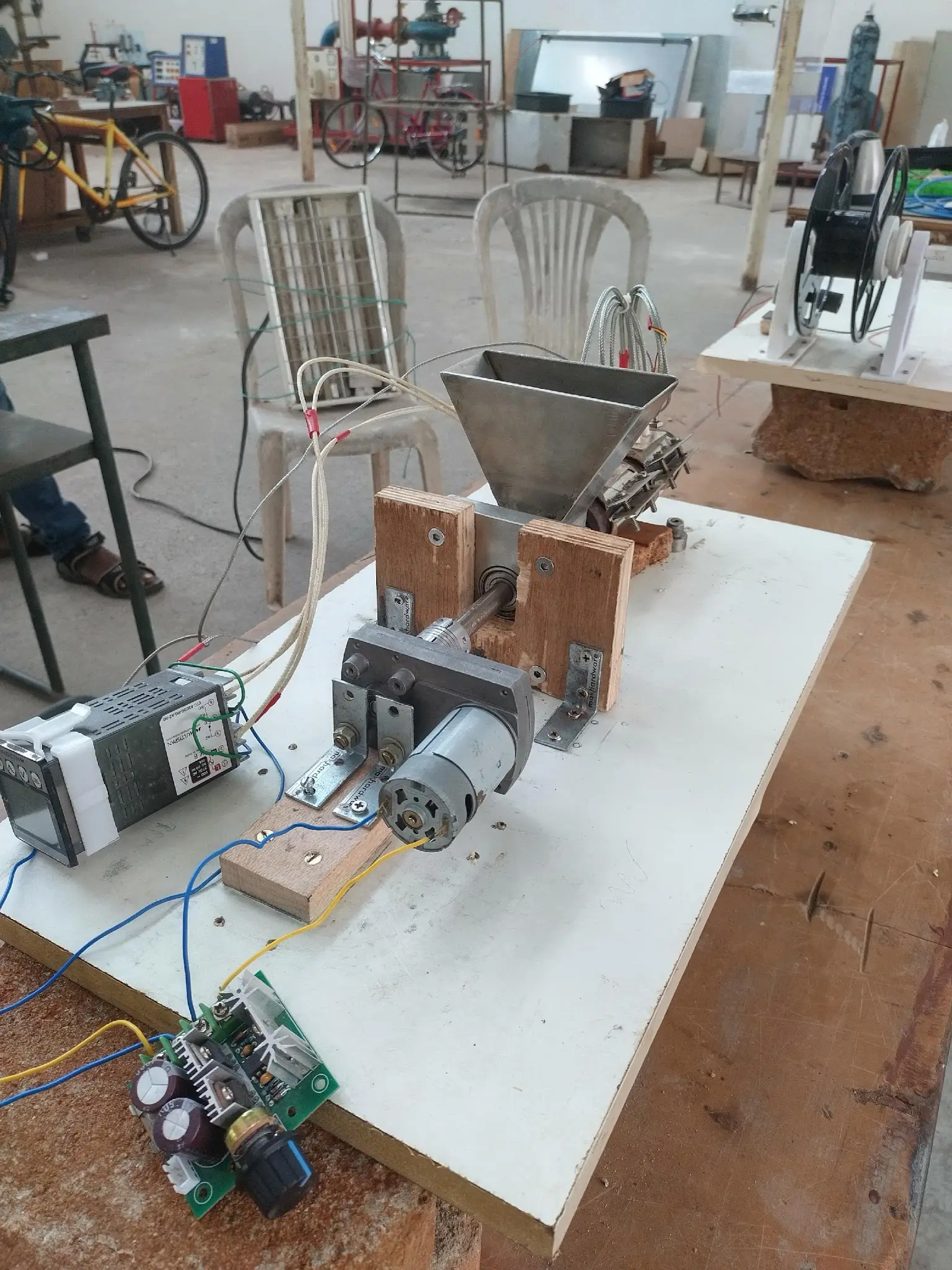 The full extruder rig on a workbench: hopper, heated barrel and screw, temperature controller, and a spool winder in the background.